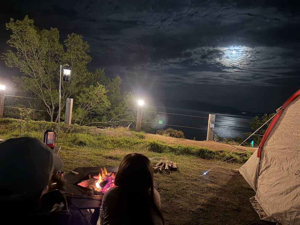 海沿いのキャンプ場の夜。焚き火の向こうに月明かりで光る海が見える